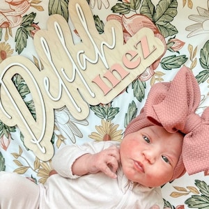 A baby girl wearing a pink bow headband and a white onesie is lying on a floral blanket. The blanket has a wooden sign with the names "Delilah Inez" written in white and pink lettering.