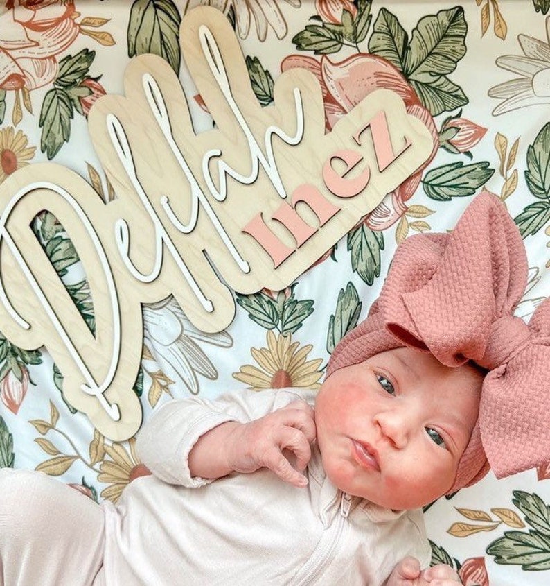 A baby girl wearing a pink bow headband and a white onesie is lying on a floral blanket. The blanket has a wooden sign with the names "Delilah Inez" written in white and pink lettering.
