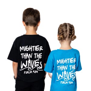 Op de afbeelding: Twee kinderen dragen bijpassende t-shirts met de tekst "Mightier Than The Waves" en Psalm 93:4. Eén shirt is zwart, de andere blauw. De kinderen zijn van achteren te zien.