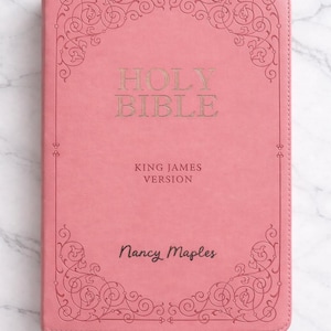 Pu&ograve; includere: Bibbia rosa con scritte dorate e decorazioni angolari. La copertina recita "Holy Bible, King James Version" e "Nancy Maples". Il libro &egrave; ambientato su uno sfondo di marmo.