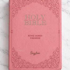 Pu&ograve; includere: Una Bibbia rosa con il testo "Holy Bible" e "King James Version" in lettere dorate. La copertina presenta un bordo ornato e il nome "Saydee" in basso.
