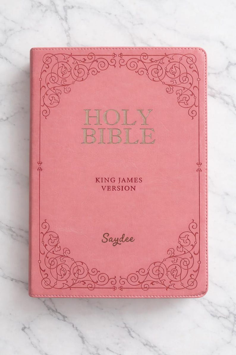 Pu&ograve; includere: Una Bibbia rosa con il testo "Holy Bible" e "King James Version" in lettere dorate. La copertina presenta un bordo ornato e il nome "Saydee" in basso.