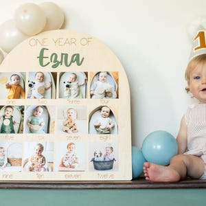 K&ouml;nnte beinhalten: Ein h&ouml;lzernes Foto-Display mit dem Text "ONE YEAR OF Ezra" und zw&ouml;lf Fotoschlitzen, jeweils mit einem Babyfoto. Ein Baby mit einer Partyh&uuml;tchen mit der Nummer "1" sitzt in der N&auml;he. Ballons in Wei&szlig; und Blau sind im Hintergrund.