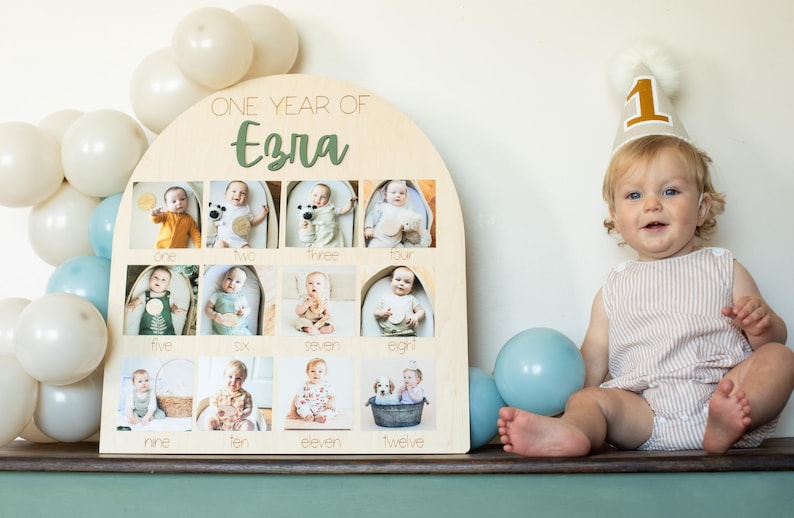 K&ouml;nnte beinhalten: Ein h&ouml;lzernes Foto-Display mit dem Text "ONE YEAR OF Ezra" und zw&ouml;lf Fotoschlitzen, jeweils mit einem Babyfoto. Ein Baby mit einer Partyh&uuml;tchen mit der Nummer "1" sitzt in der N&auml;he. Ballons in Wei&szlig; und Blau sind im Hintergrund.