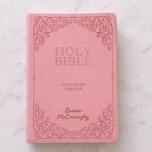 May include: A pink Holy Bible, King James Version, with decorative corner accents. The cover features the words "HOLY BIBLE" and the author's name, "Bonnie McConaughy."