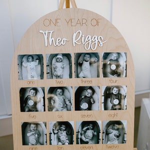 K&ouml;nnte beinhalten: Holz-Foto-Display mit zw&ouml;lf Schwarz-Wei&szlig;-Babyfotos, eines f&uuml;r jeden Monat des ersten Jahres. Das Display ist oben gew&ouml;lbt und enth&auml;lt den Text "ONE YEAR OF Theo Riggs". Das Display ist auf einer Holzstaffelei montiert.