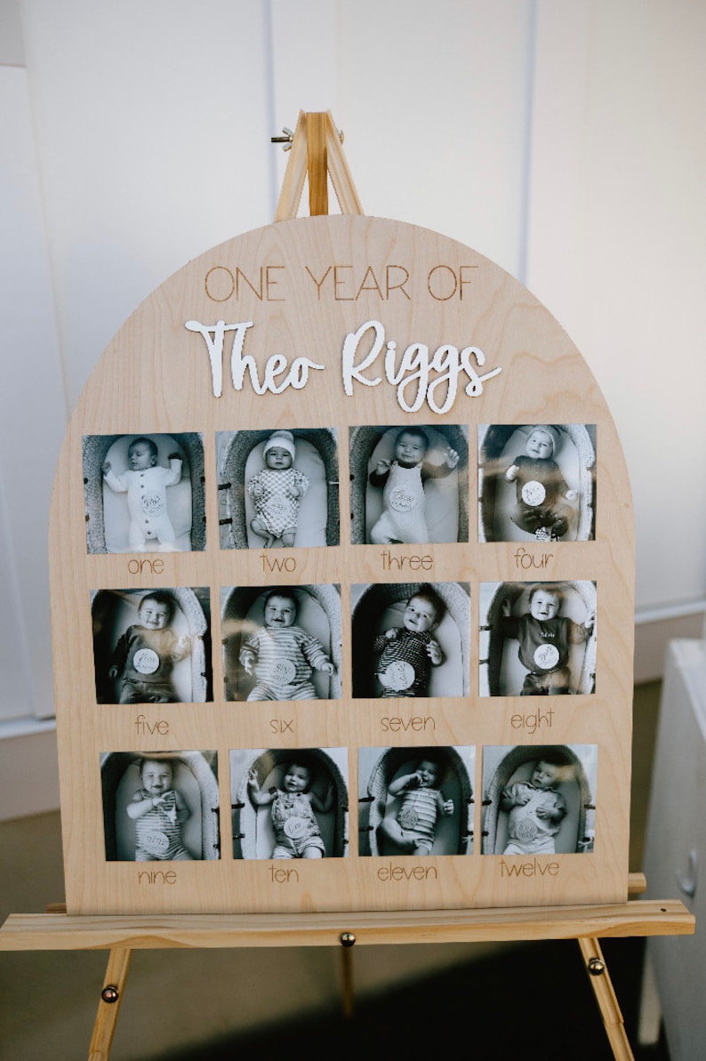 K&ouml;nnte beinhalten: Holz-Foto-Display mit zw&ouml;lf Schwarz-Wei&szlig;-Babyfotos, eines f&uuml;r jeden Monat des ersten Jahres. Das Display ist oben gew&ouml;lbt und enth&auml;lt den Text "ONE YEAR OF Theo Riggs". Das Display ist auf einer Holzstaffelei montiert.