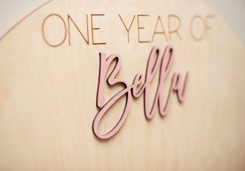K&ouml;nnte beinhalten: Ein Holzschild mit den Worten "ONE YEAR OF" und dem Namen "Bella" in einer rosa Schreibschrift. Der Name ist rosa, und das Schild ist aus hellem Holz gefertigt.