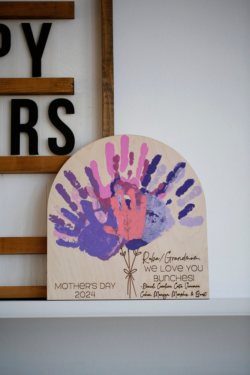 A wooden arch with handprints in shades of pink, purple, and blue. The arch is decorated with a floral design and the text "Reba/Grandmom. We LOVE YOU BUNCHESI -Daniel Carolina Cata Vivianan Cohen Mansyon Murphus & Quest" and "MOTHER'S DAY 2024".