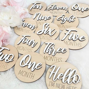 May include: A set of twelve wooden baby milestone discs with white text. The discs feature the words "One Year", "Eleven Months", "Ten Months", "Nine Months", "Eight Months", "Seven Months", "Six Months", "Five Months", "Four Months", "Three Months", "Two Months", "One Month", and "Hello I'm New Here".
