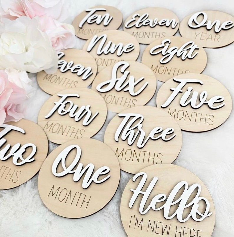 May include: A set of twelve wooden baby milestone discs with white text. The discs feature the words "One Year", "Eleven Months", "Ten Months", "Nine Months", "Eight Months", "Seven Months", "Six Months", "Five Months", "Four Months", "Three Months", "Two Months", "One Month", and "Hello I'm New Here".