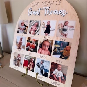 K&ouml;nnte beinhalten: Eine h&ouml;lzerne Foto-Anzeigetafel mit gew&ouml;lbtem Oberteil, die eine Sammlung von zw&ouml;lf Babyfotos zeigt. Die Tafel ist mit dem Text "ONE YEAR OF Grant Thomas" graviert und enth&auml;lt Metallst&uuml;tzbeine.