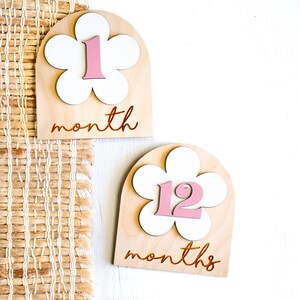 Two wooden monthly milestone markers with a white flower design and pink numbers. The first marker says "1 month" and the second marker says "12 months".