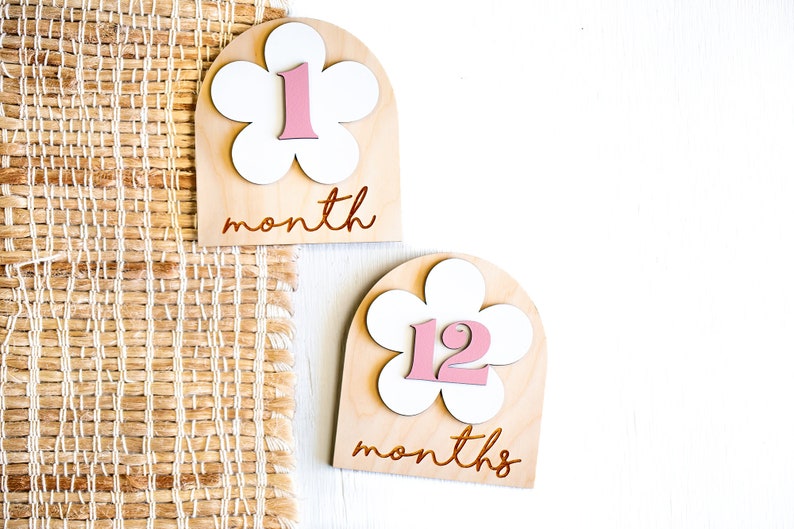Two wooden monthly milestone markers with a white flower design and pink numbers. The first marker says "1 month" and the second marker says "12 months".