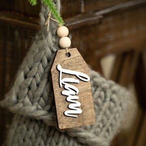 May include: A wooden stocking tag with the name "Liam" written in white lettering. The tag is attached to a gray knitted stocking with two wooden beads.