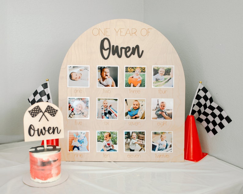 K&ouml;nnte beinhalten: Eine h&ouml;lzerne Fototafel mit dem Text "ONE YEAR OF Owen" und zw&ouml;lf Fotorahmen. Die Tafel ist mit einem kleinen Kuchen, Schachflaggen und einem roten Verkehrskegel dekoriert. Jeder Fotorahmen ist mit einer Zahl beschriftet.