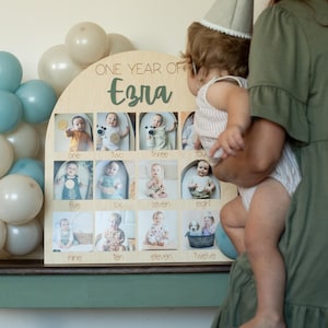 K&ouml;nnte beinhalten: Ein h&ouml;lzernes Foto-Display mit dem Text "ONE YEAR OF Ezra" und zw&ouml;lf Fotoschlitzen. Das Display ist mit Fotos eines Babys dekoriert. Ballons in Blau- und Wei&szlig;t&ouml;nen befinden sich im Hintergrund.