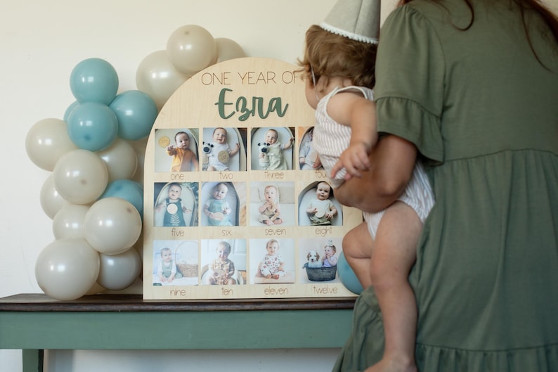 K&ouml;nnte beinhalten: Ein h&ouml;lzernes Foto-Display mit dem Text "ONE YEAR OF Ezra" und zw&ouml;lf Fotoschlitzen. Das Display ist mit Fotos eines Babys dekoriert. Ballons in Blau- und Wei&szlig;t&ouml;nen befinden sich im Hintergrund.