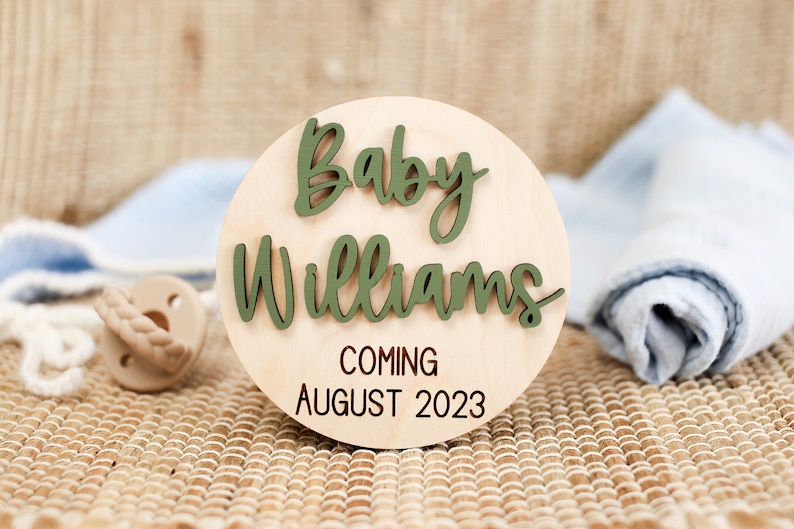 May include: A round wooden sign with the text "Baby Williams Coming August 2023". The words "Baby Williams" are in green raised letters.