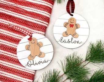 May include: Two white round wooden ornaments with gingerbread people cutouts. The ornaments are decorated with red and white striped ribbon and the names "Olivia" and "Easton" are written on them.