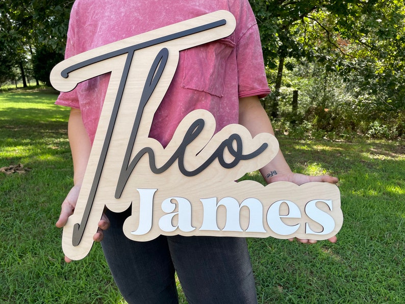 A wooden sign with the name "Theo James" written in black and white lettering. The sign is held by a person wearing a pink shirt.