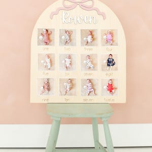 May include: A wooden monthly milestone board with a light pink bow and the name 'Rowen' in cursive. The board has twelve square openings, each with a baby photo and the corresponding month number written below. The board is displayed on a light green stool against a pink background.