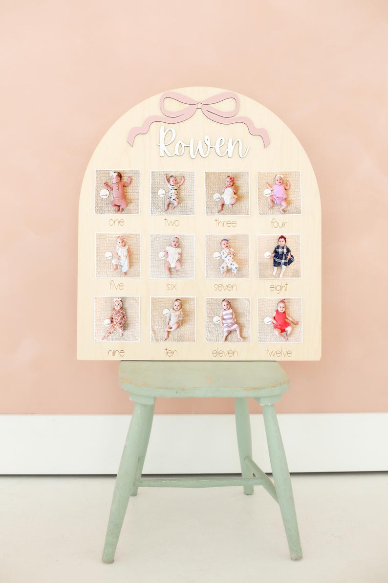May include: A wooden monthly milestone board with a light pink bow and the name 'Rowen' in cursive. The board has twelve square openings, each with a baby photo and the corresponding month number written below. The board is displayed on a light green stool against a pink background.
