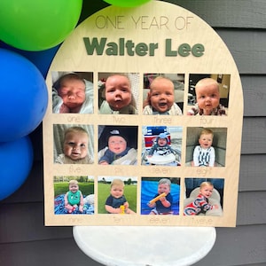 K&ouml;nnte beinhalten: Eine Holztafel mit der Aufschrift "ONE YEAR OF Walter Lee" und zw&ouml;lf kleinen Fotos eines Babys, die jeweils mit einer Monatsnummer beschriftet sind. Gr&uuml;ne und blaue Luftballons sind im Hintergrund zu sehen.
