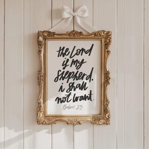 May include: A framed print with the quote "The Lord is my shepherd. I shall not want." in black script. The ornate gold frame is hung with a white ribbon and bow against a white wood-paneled wall. Psalm 23 is written at the bottom.