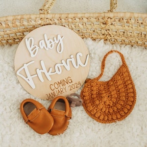 May include: A wooden sign with the text "Baby Frkovic Coming January 2024" in white lettering. The sign is surrounded by a brown crocheted bib, a pair of brown baby shoes, and a brown dummy.