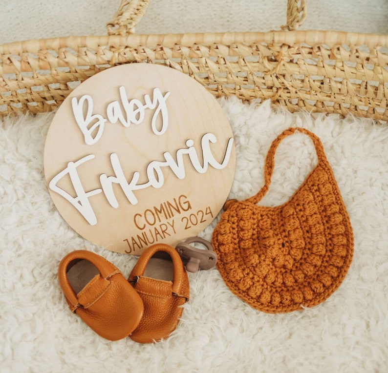 May include: A wooden sign with the text "Baby Frkovic Coming January 2024" in white lettering. The sign is surrounded by a brown crocheted bib, a pair of brown baby shoes, and a brown dummy.