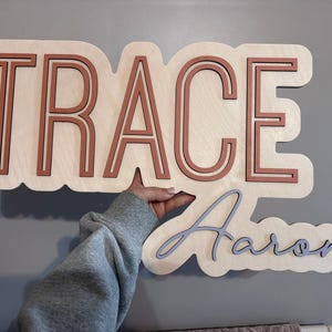 Wooden nursery name sign "Trace Aaron" in orange and gray on a natural wood