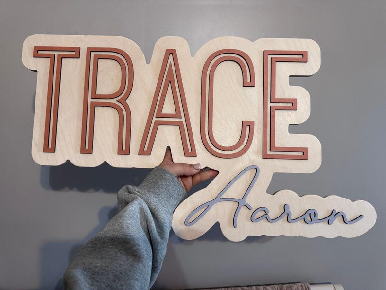 Wooden nursery name sign "Trace Aaron" in orange and gray on a natural wood