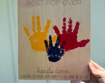 May include: Wooden sign with the text "Best Pop Ever" and handprints in yellow, red, and blue paint. The text "hands down" and "Love, Charlee Beth, Silas, & Evie" is also on the sign.