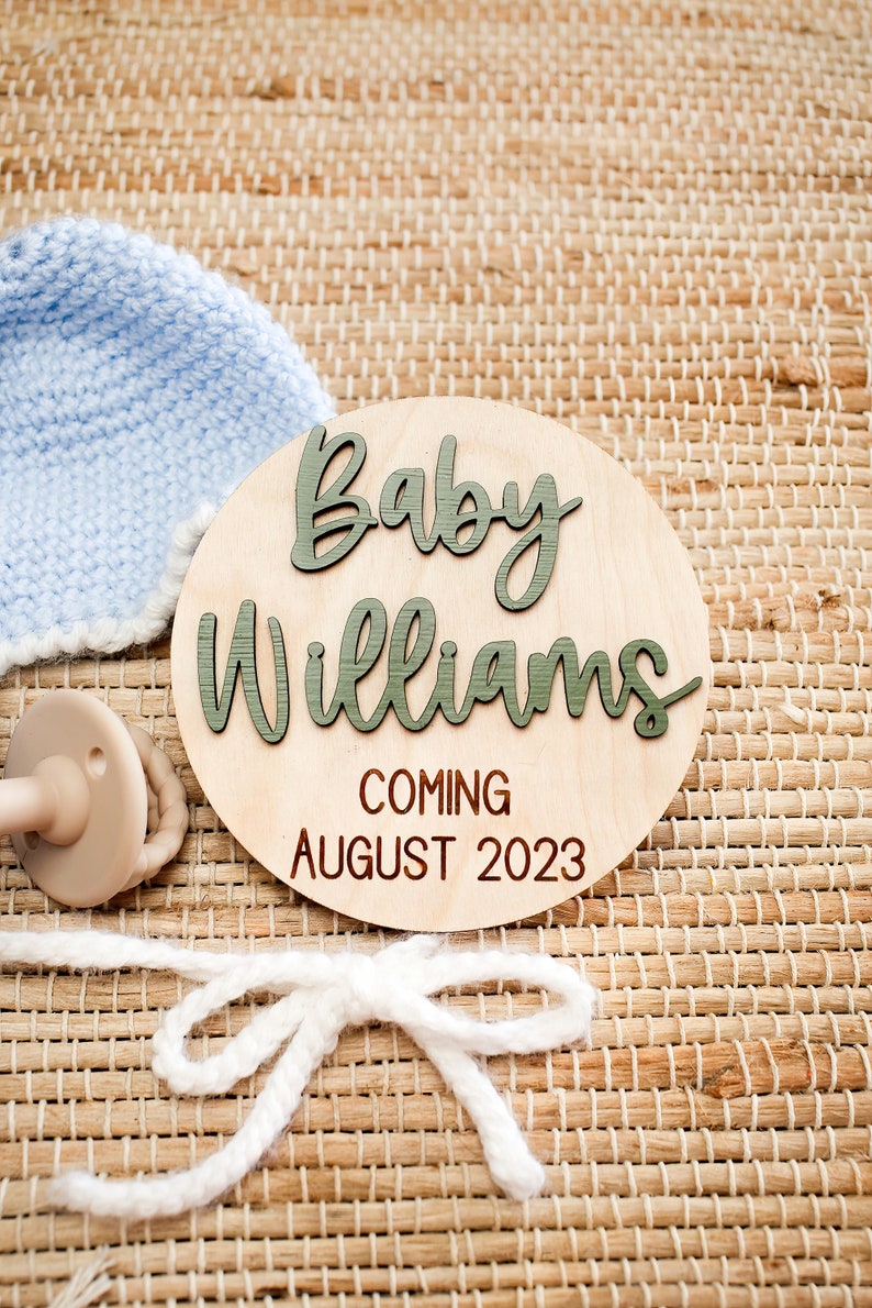 May include: A wooden baby announcement sign with the text "Baby Williams Coming August 2023". The sign is decorated with a blue knitted hat and a beige dummy.