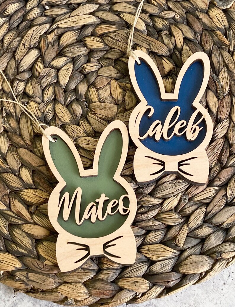 May include: Two wooden bunny-shaped ornaments with names. One is green with the name "Mateo" and the other is blue with the name "Caleb". Both have a bow tie design and are hanging by twine strings. The background is a woven, brown surface.