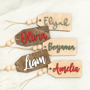 May include: Five wooden gift tags with names written on them in different colors. The names are Elijah, Olivia, Benjamin, Liam, and Amelia. Each tag has a string attached to it with wooden beads.