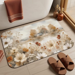 Op de afbeelding: Een rechthoekige badmat met een aquarel-landschapsprint van een klein huis bij een waterpartij, omringd door witte, crèmekleurige en oranje wilde bloemen. De mat heeft afgeronde hoeken en een zacht, gedempt kleurenpalet van aardetinten.