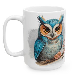 May include: White ceramic mug featuring a detailed illustration of a blue and orange owl perched on a branch with pink flowers. The owl has large, expressive eyes and is set against a soft gray background. The mug has a large handle.