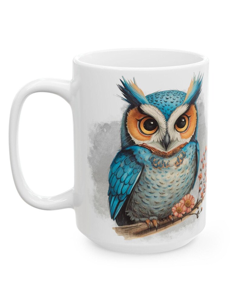 May include: White ceramic mug featuring a detailed illustration of a blue and orange owl perched on a branch with pink flowers. The owl has large, expressive eyes and is set against a soft gray background. The mug has a large handle.
