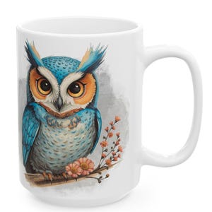 May include: White ceramic mug featuring a detailed illustration of a blue and orange owl perched on a branch with pink flowers. The owl has large, expressive eyes and is set against a soft gray background.