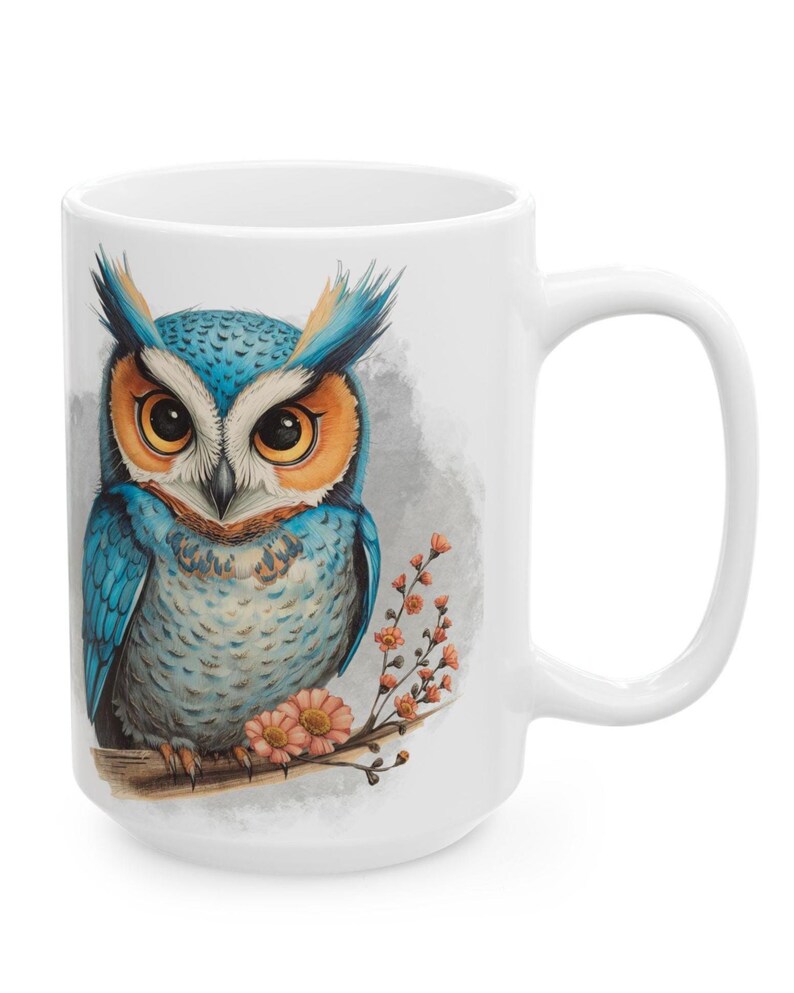 May include: White ceramic mug featuring a detailed illustration of a blue and orange owl perched on a branch with pink flowers. The owl has large, expressive eyes and is set against a soft gray background.
