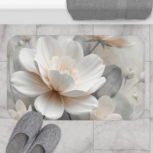 May include: A rectangular bath mat featuring a large, white floral design with soft peach and gray accents. The mat is placed on a tiled floor, with a pair of gray slippers and a gray towel visible.