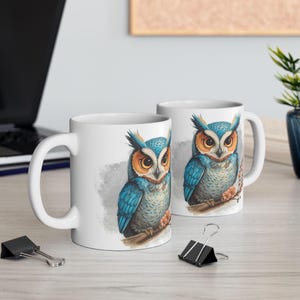 May include: Two white ceramic mugs with a detailed illustration of a blue and orange owl perched on a branch. The owl has intricate feather details and is set against a soft, watercolor-style background. The mugs are on a light-colored surface.