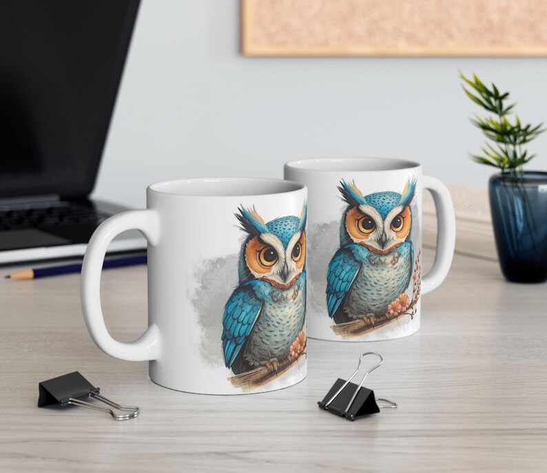 May include: Two white ceramic mugs with a detailed illustration of a blue and orange owl perched on a branch. The owl has intricate feather details and is set against a soft, watercolor-style background. The mugs are on a light-colored surface.