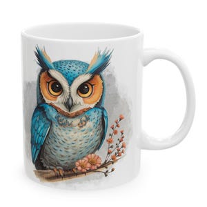 May include: White ceramic mug featuring a detailed illustration of a blue and orange owl perched on a branch with small orange flowers. The owl has large, expressive eyes and is set against a soft, gray background. The mug has a curved handle.
