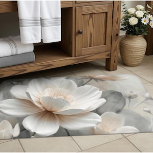 May include: A rectangular bath mat featuring a large, central white and pale peach lotus flower graphic. The design includes soft gray and white watercolor-style background elements with smaller flower buds, creating a floral pattern on the mat.