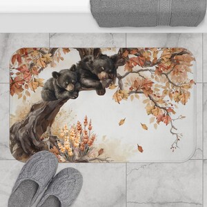 Op de afbeelding: Een rechthoekige badmat met een aquarelillustratie van twee zwarte berenwelpen die op een boomtak rusten. De scène bevat herfstbladeren in tinten oranje, bruin en geel, met een groepje wilde bloemen aan de onderkant van de afbeelding.