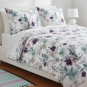 May include: A white bed with a floral patterned comforter and two matching pillowcases. The floral design features purple, teal, and gray flowers and leaves. A white lamp sits on a nightstand next to the bed.