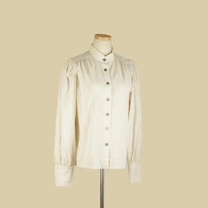 May include: A cream-colored long-sleeve blouse with a high collar and button-down front. The blouse features subtle pleating at the shoulders and cuffs. The garment is displayed on a mannequin, showcasing its tailored fit and classic design.